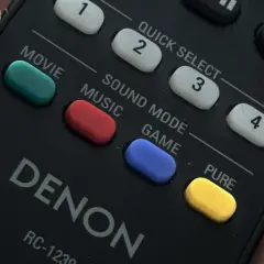 Dłoń trzyma pilot Denon. Widoczne przyciski: Movie, Music, Game, a także żółty przycisk PURE.