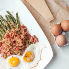 Zielone szparagi z chrupiącym boczkiem i jajka sadzone. Obok kawałek parmezanu i jajka w wytłoczce.