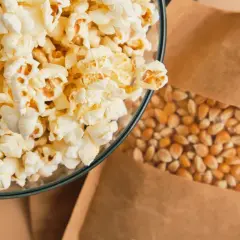 Miseczka pełna puszystego popcornu obok ziaren kukurydzy w papierowej torbie. Czy popcorn jest lekkostrawny? Zależy od dodatków!