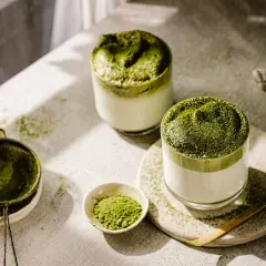 Zwei Gläser cremiger Matcha-Latte, bestreut mit feinem Pulver. Die Zubereitung mit Chasen und Sieb deutet auf die belebende matcha wirkung hin.