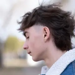 Junger Mann mit modernen Mullets, der nachdenklich zur Seite blickt. Sein Haar ist dunkelbraun und zerzaust, die Seiten sind kurz geschoren. Er trägt eine Jeansjacke mit Teddyfellkragen.