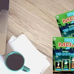 Dwie zdrapki lotto "Moje 7" z napisem "Wygraj 75000zł" leżą obok laptopa i kubka kawy.