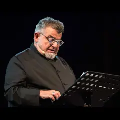 Giancarlo Cauteruccio, con occhiali e barba, legge da un leggio su un palco buio.