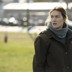 Detektyw z "Mare of Easttown" w brązowej kurtce i szarym szaliku, z czerwoną wstążką na piersi, stoi na tle zielonej łąki i samochodów.