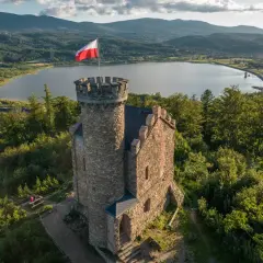 Kamienny zamek henryka z polską flagą na wieży, otoczony zielenią, z jeziorem i górami w tle.