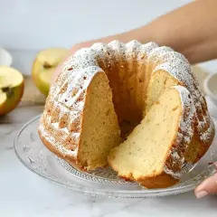 Saftiger Gugelhupf mit Puderzucker bestäubt, daneben Äpfel und Apfelmus. Ein Stück ist bereits angeschnitten.