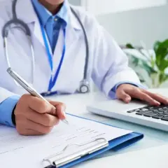 Lekarz w białym kitlu z długopisem w dłoni i stetoskopem na szyi, pisze dokumenty, pracując przy laptopie.