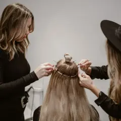 Två personer hjälper till att **slinga håret hemma** med extensions. En med svart polotröja och en med svart hatt arbetar på det långa blonda håret.