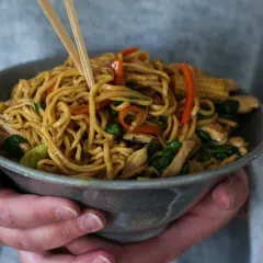 En skål med lo mein-nudlar, kyckling och grönsaker, lyfts med ätpinnar.