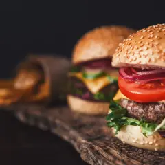 Dwa domowe burgery z serem, pomidorem i cebulą na drewnianej desce, obok frytki.