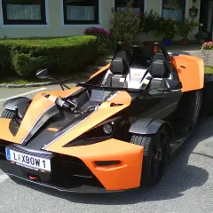 Pomarańczowy KTM X-Bow z czarnymi akcentami i tablicą rejestracyjną "XBOW 1" stoi na parkingu przed budynkiem z zielonym żywopłotem.