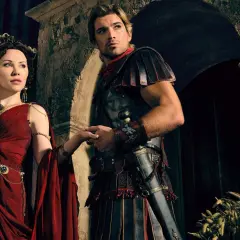 Die Besetzung von Spartacus: Ashur und Lucretia, in prachtvollen Gewändern und Rüstungen, halten Händchen vor einer antiken Kulisse.