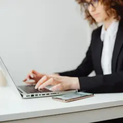 Kobieta w okularach i czarnej marynarce pracuje na laptopie. Obok leży telefon. Może szuka informacji, co to jest upadłość konsumencka.
