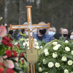 Drewniany krzyż z Jezusem, otoczony wieńcami, symbolizuje pożegnanie. Po pogrzebie tabliczka z napisem "Spoczywaj w pokoju" pozostaje jako pamiątka.