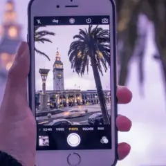 Dłoń trzyma iPhone'a, na ekranie widać budynek z zegarem i palmy. Dowiedz się, jak ustawić siatkę w aparacie iPhone, by ułatwić kompozycję.