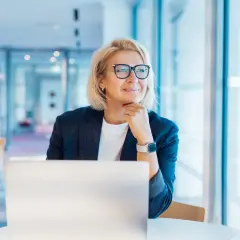 Donna bionda con occhiali, seduta al computer, pensa al suo futuro. Un nuovo capitolo, cambiare lavoro a 50 anni, è una possibilità concreta.