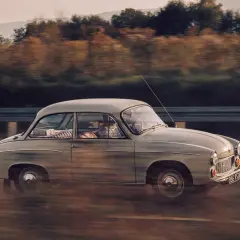 Klasyczny samochód w ruchu, uchwycony techniką **panning**, mija jesienny krajobraz.