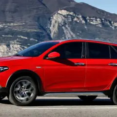 Czerwony Fiat Tipo Cross, wszechstronny samochód z segmentu kombi, przemierza malowniczą drogę nad jeziorem, z górami w tle.