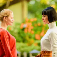 Due donne si guardano intensamente, una con i capelli biondi raccolti, l'altra con un caschetto scuro. Si percepisce un'atmosfera di invidia e gelosia tra loro.