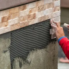 Close-up of a worker's hands carefully performing wall tile installation, applying a new stone tile to a textured adhesive layer.