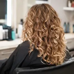 Kobieta w czarnym szlafroku, z pięknymi, złotymi loki trwała. W tle widać salon fryzjerski.