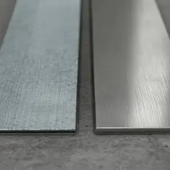 Dwa metalowe paski leżą obok siebie na betonowym podłożu. Jeden jest szorstki, drugi gładki, jakby przygotowany do polerowania.