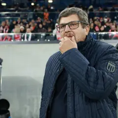 Włoski trener w okularach, zamyślony, w granatowej kurtce z naszywką "ITALIA", stoi na tle trybun stadionu.