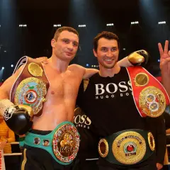 Two boxers, one with multiple championship belts, stand together. The boxer on the left, Vitali Klitschko, shows his impressive height and championship status.