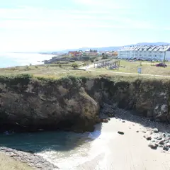 Acantilados rocosos y una pequeña cala de arena en la playa de San Bartolo Barreiros, con edificios residenciales al fondo.