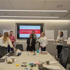 A team coach leads a meeting in a conference room, with a screen displaying "What will make today successful for you?".
