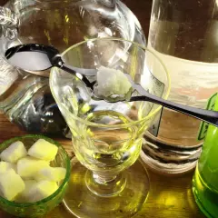 Tradycyjny sposób na to, jak pić absynt: kostka cukru na łyżeczce nad kieliszkiem, obok dzbanek z wodą i butelka z zielonym alkoholem.