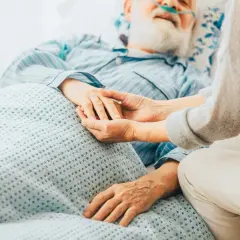 Ein älterer Mann mit Bart und Nasenbrille liegt im Bett. Eine Person hält seine Hand. Die Szene vermittelt die Endphase, wie man an Prostatakrebs stirbt.