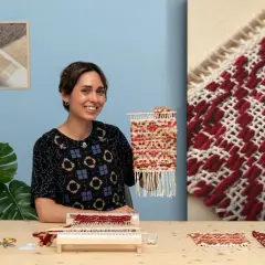 Artisanat textile : une femme souriante tient une petite tapisserie colorée. Sur la table, du fil, des ciseaux et un métier à tisser.