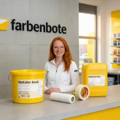 Frau mit roten Haaren lächelt, neben Farbeimer, Rolle und Klebeband. Alles, was man zum Streichen braucht, ist hier zu sehen.