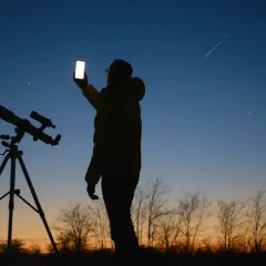 Sylwetka osoby z telefonem i teleskopem na tle nieba z zaćmieniem Księżyca i smugą po samolocie.