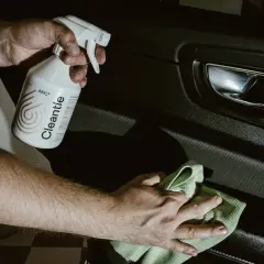 Ręce czyszczą wnętrze samochodu preparatem APC. Butelka z etykietą "Cleantle APC²" i zielona mikrofibra.