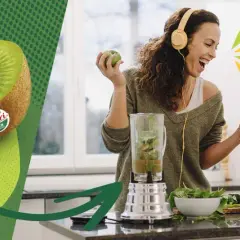 Glad kvinna med hörlurar gör smoothie med kiwi nyttigt. Två halverade kiwifrukter syns till vänster.