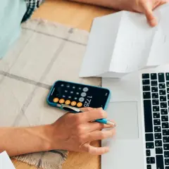 Osoba analizuje dokumenty, korzystając z kalkulatora w telefonie i laptopa, by obliczyć maksymalne oprocentowanie kredytu.