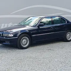 Ciemnoniebieskie BMW 7 E38 z lat 90. stoi na żwirowym podjeździe, z odbiciem chmur w szybach.
