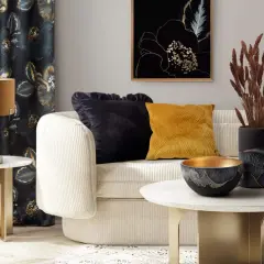 Elegancki salon z kanapą, ozdobnymi poduszkami i wazonem z suszonymi trawami zamiast kwiatów. Złote akcenty i ciemne zasłony tworzą przytulną atmosferę.