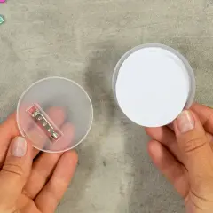 Zwei Hände halten Teile für das **button basteln**: eine mit einer transparenten Rückseite und Nadel, die andere mit einem weißen Kreis.