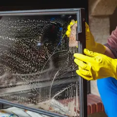 Mit gelben Handschuhen wird das Kaminglas gereinigt. Schaum und Schlieren bedecken die Scheibe, während eine Hand mit einem Schwamm schrubbt.