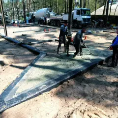 Pracownicy wylewają beton na budowie, przygotowując podłoże pod posadzkę. Widać ciężarówkę z mieszanką betonową i sprzęt budowlany.