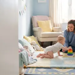 Uśmiechnięta mama patrzy, jak niemowlę odpycha się do tyłu na plecach, bawiąc się kolorową zabawką na dywanie w pokoju dziecięcym.