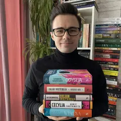 Mężczyzna w okularach trzyma stos książek, wśród nich "Odyseja" i dzieła Colsona Whiteheada. W tle regały pełne literatury, przypominające o bogactwie polskiej i światowej prozy, jak u Bolesława Prusa.