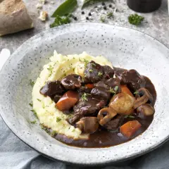 En mustig boeuf bourguignon med potatismos, champinjoner och morötter. Serveras i en rustik skål med en bit bröd och ett glas vatten.