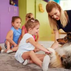 Uśmiechnięta dziewczynka głaszcze psa terapeutycznego, siedząc w grupie dzieci.