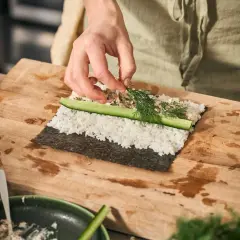 Händer lägger dill på egen sushi med ris, gurka och nori.