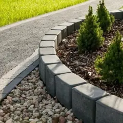 Obrzeże rabaty z kostki brukowej, wyznaczające granicę między ścieżką a zielenią. Kamienie i młode drzewka dopełniają aranżacji.