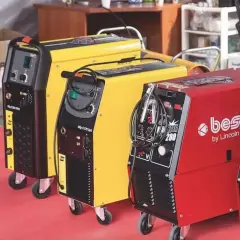 Trzy spawarki: czarna, żółta i czerwona, gotowe do pracy. Każda z nich ma uchwyt i kable, a czerwona posiada napis "best by Lincoln Electric".